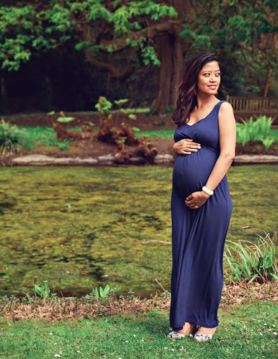 Ania Chandra maternity Photographer in Berkshire. Photo of pregnant women holding her belly with her baby inside and smiling. Background is very green and beautiful pond behind her in Harris Garden Reading.