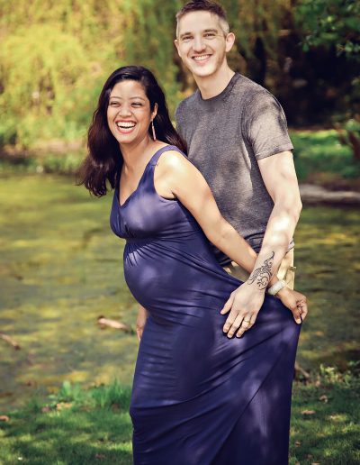 Ania Chandra Maternity Photographer in Berkshire and beyond. Pregnant mother with her husband standing and laughing together in beautiful nature garden, green background with pond.