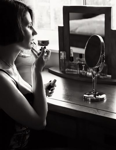 boudoir by ania chandra in reading area, women sitting infront of the mirror and doing her makeup - black and white image - she looks very beautiful she also has short her up to her ears and she is wearing only body suit