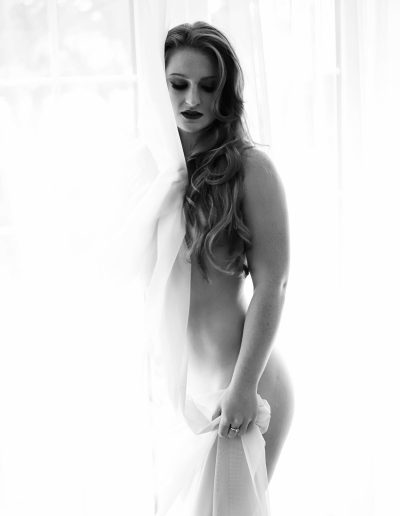 naked women is standing next to he window and her hair are covering her breast and she is covered holding white curtain. Ania chandra photographer in Reading - Boudoir image