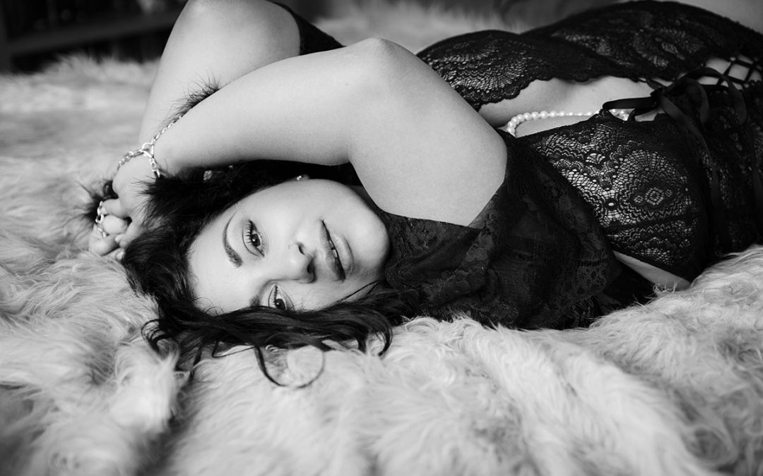 Curvie women with black hair and beautiful big eyes is lying on the bed with hands up and facing camera - smiling ever so gently - Boudoir photographer Reading -Ania chandra photographer