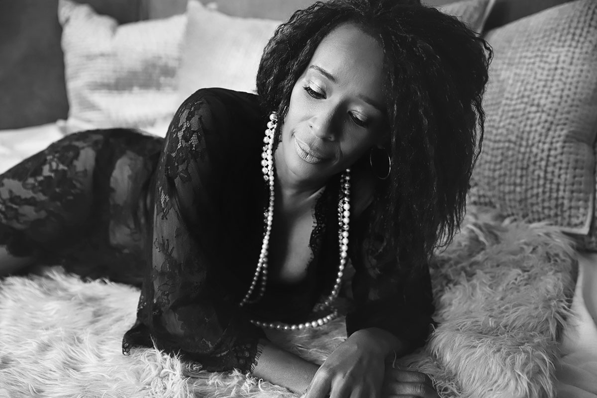 Beautiful black women is lying on her front looking down - dressed in black gown and purl neckless on her neck - image done by Boudoir Ania Chandra Photography in Rearing 