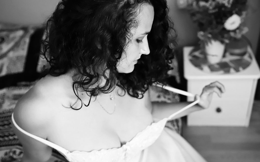 black and white image of women in her wedding dress but she is holding off to her strap and showly pulling it away - iboudoir image done by ania chandra photographer in Reading